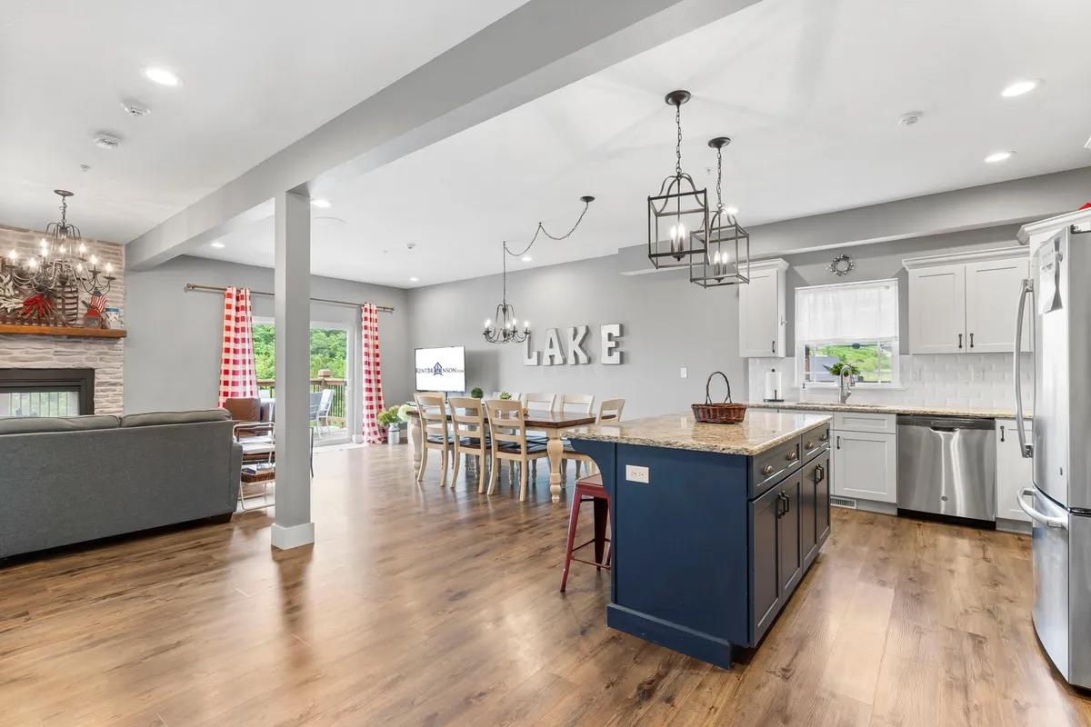 This image showcases a warm, open-concept main area with a kitchen island, a spacious dining table, a cozy fireplace, and a glimpse of the lake through large windows. Decorative accents and comfortable seating further amplify the inviting communal atmosphere.