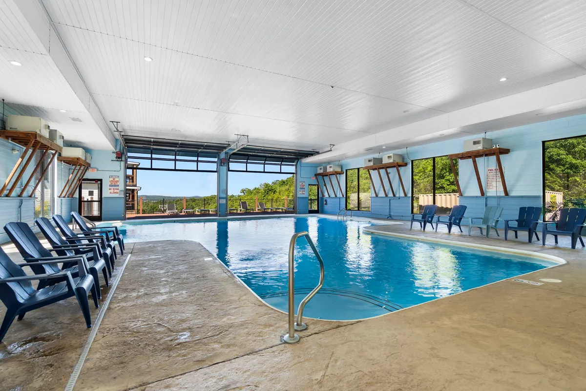 A serene interior scene of the indoor pool, highlighted by expansive windows that let in natural light. Lounge chairs are arranged poolside, inviting relaxation and socialization regardless of the weather outside. The tranquil water and inviting seating create a year-round retreat for guests of all ages.