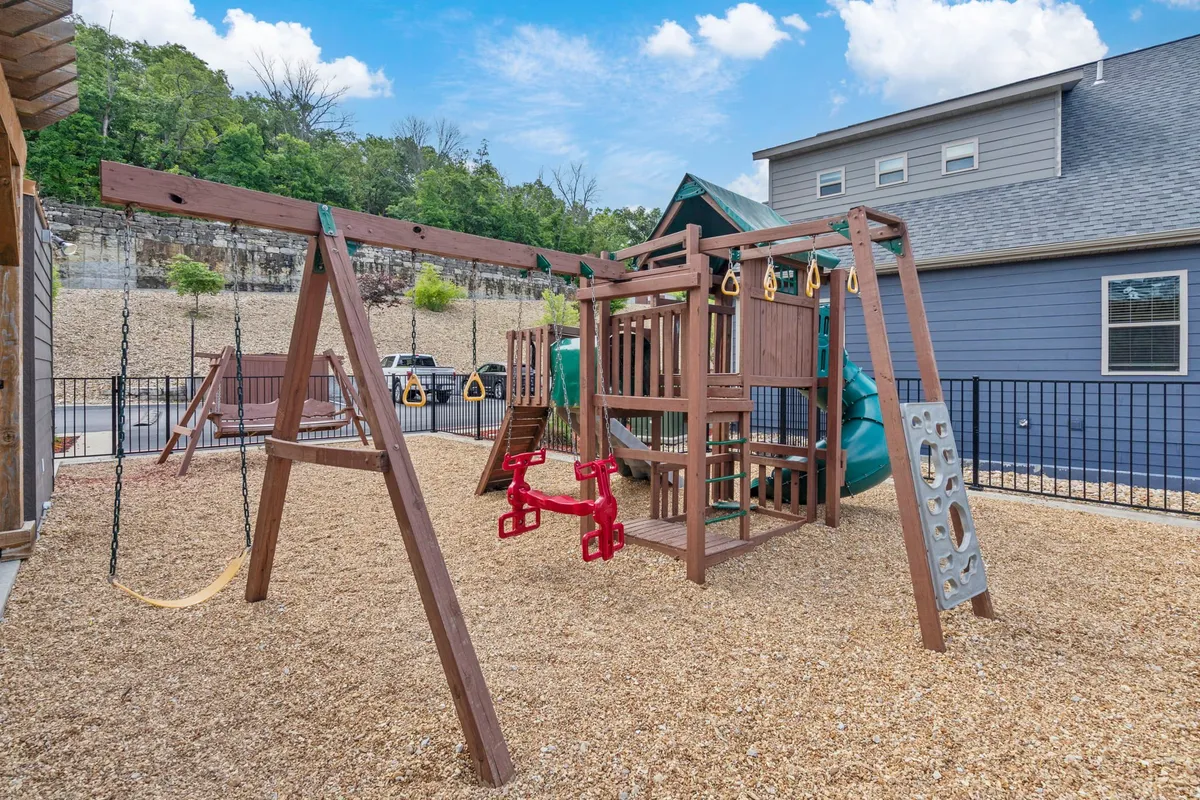A cheerful outdoor playground area with a swing set, slide, and gravel flooring, surrounded by green landscaping. Children can be seen playing, and the setup encourages outdoor activity, socializing, and joyful family moments.