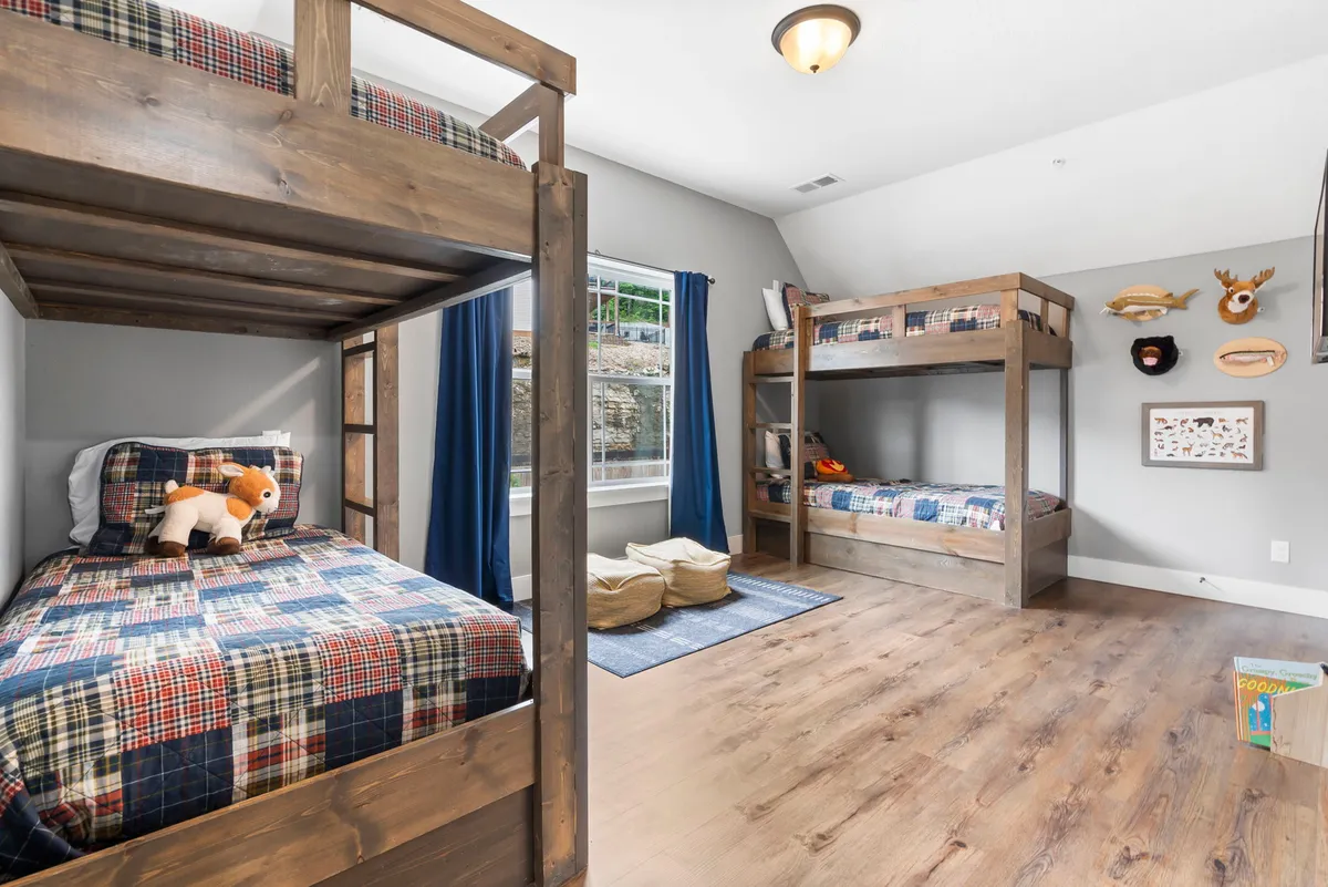 A cozy bunk room featuring bunk beds with plaid bedding, plush stuffed animals, and a large window that lets in natural light. Wall decor adds a playful touch, making the space warm and inviting for children.