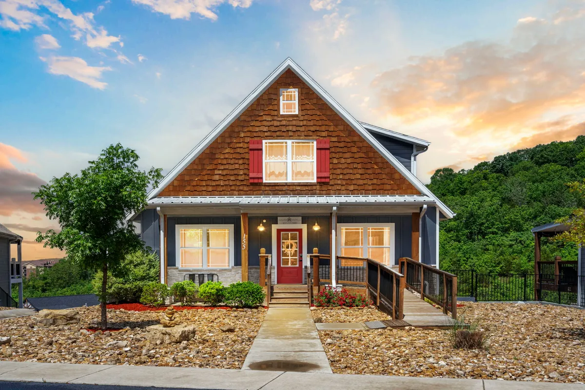 The hero image showcases the charming exterior of Hawthorn House in Branson, Missouri. A welcoming front porch with cozy seating invites relaxation, surrounded by lush landscaping and classic cottage-style windows. The unique roof design adds character, while the scene evokes the perfect setting for family gatherings, savoring sunsets, or celebrating special moments outdoors.