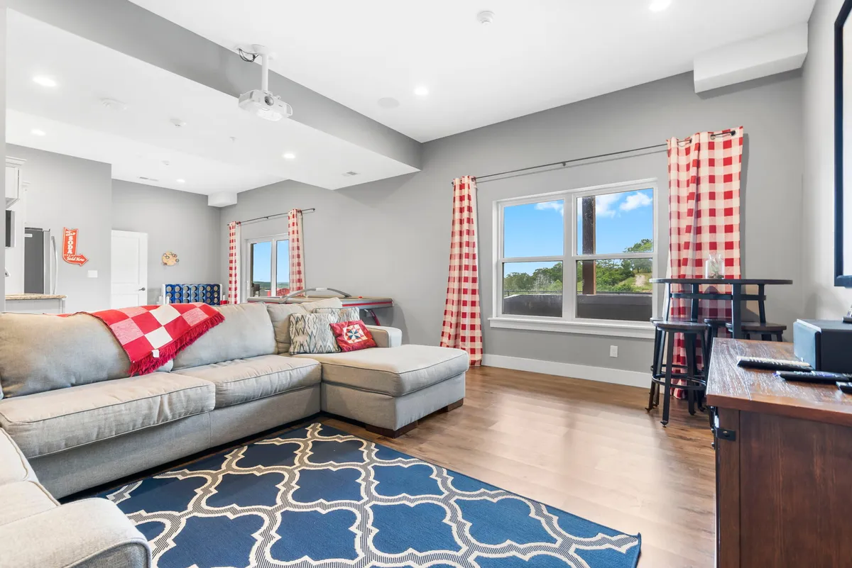 A cozy interior with a large sectional sofa, vibrant throw pillows, an area rug with a geometric pattern, and a window with checkered curtains. The inviting atmosphere is perfect for relaxing, watching movies, or gathering for social games.