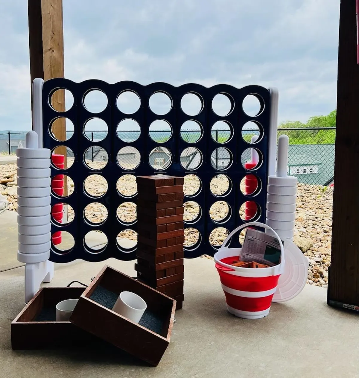 This exterior scene features a large outdoor Connect Four game, a stack of jumbo Jenga blocks, and colorful buckets with cups, all set up in a landscaped yard. The setting is lively and designed for outdoor fun, with grass underfoot and ample space for group play and social gatherings.