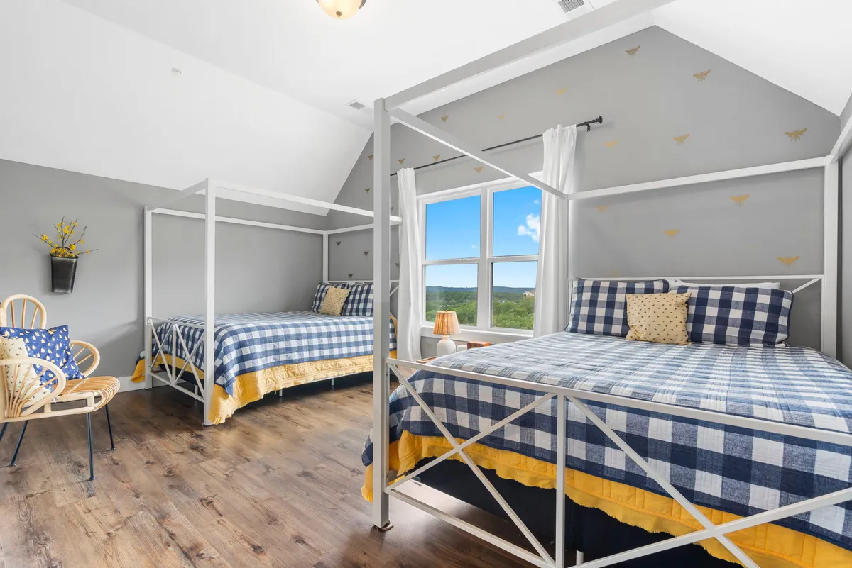A spacious bedroom with elegant canopy beds, large windows that flood the room with sunlight, wooden floors, and decorative pillows. The setting feels both upscale and welcoming, suitable for a king or double queen suite.