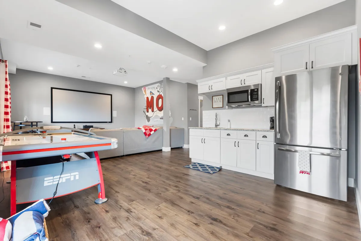 A vibrant interior showing a lower-level entertainment area with an air hockey table in the foreground, a spacious kitchenette with a full fridge, a large projection screen for movie nights, and a comfortable couch area with red and white checkered curtains. The scene is energetic, inviting, and perfect for group activities.