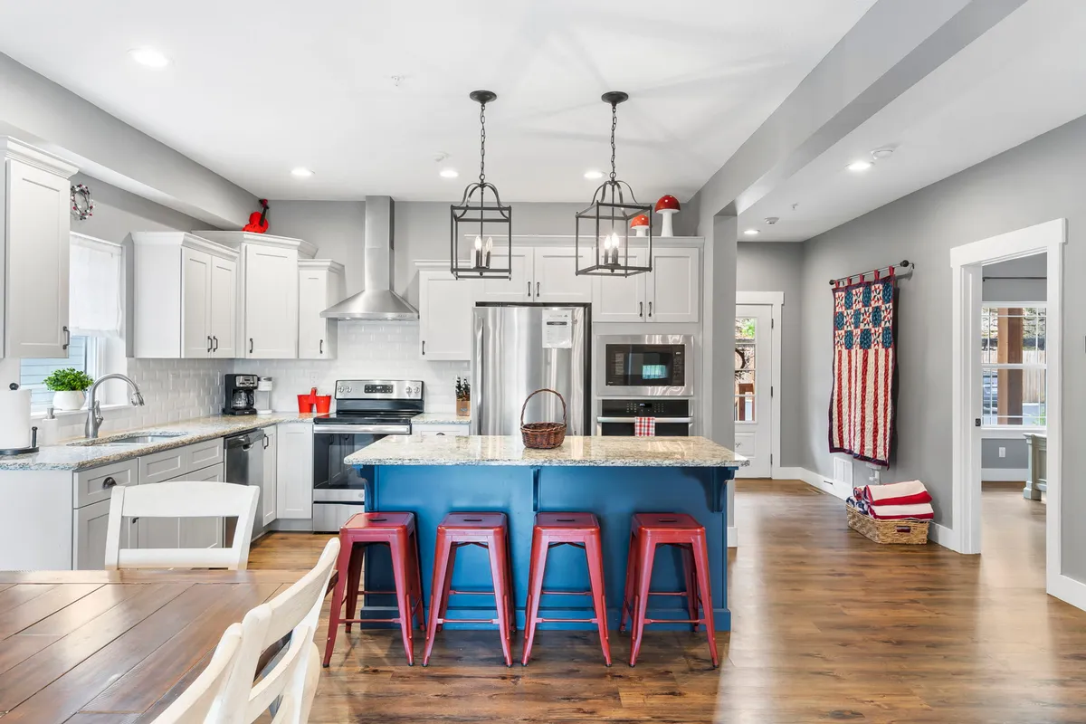 A bright, inviting kitchen with a central island, red barstools, granite countertop, modern appliances, and a textile wall hanging. The space is filled with natural light, ideal for group cooking and gatherings.