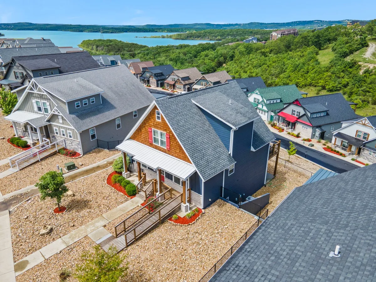 An exterior shot of the main house with the lake glistening in the background, surrounded by landscaped flowers and neighboring homes. The scene captures an atmosphere of cheerful relaxation, community, and connection to the waterfront.